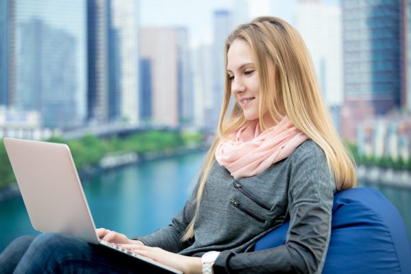Young girl using laptop, study, working, communicating, ordering online, sitting in comfortable beanbag chair, cityscape on the background